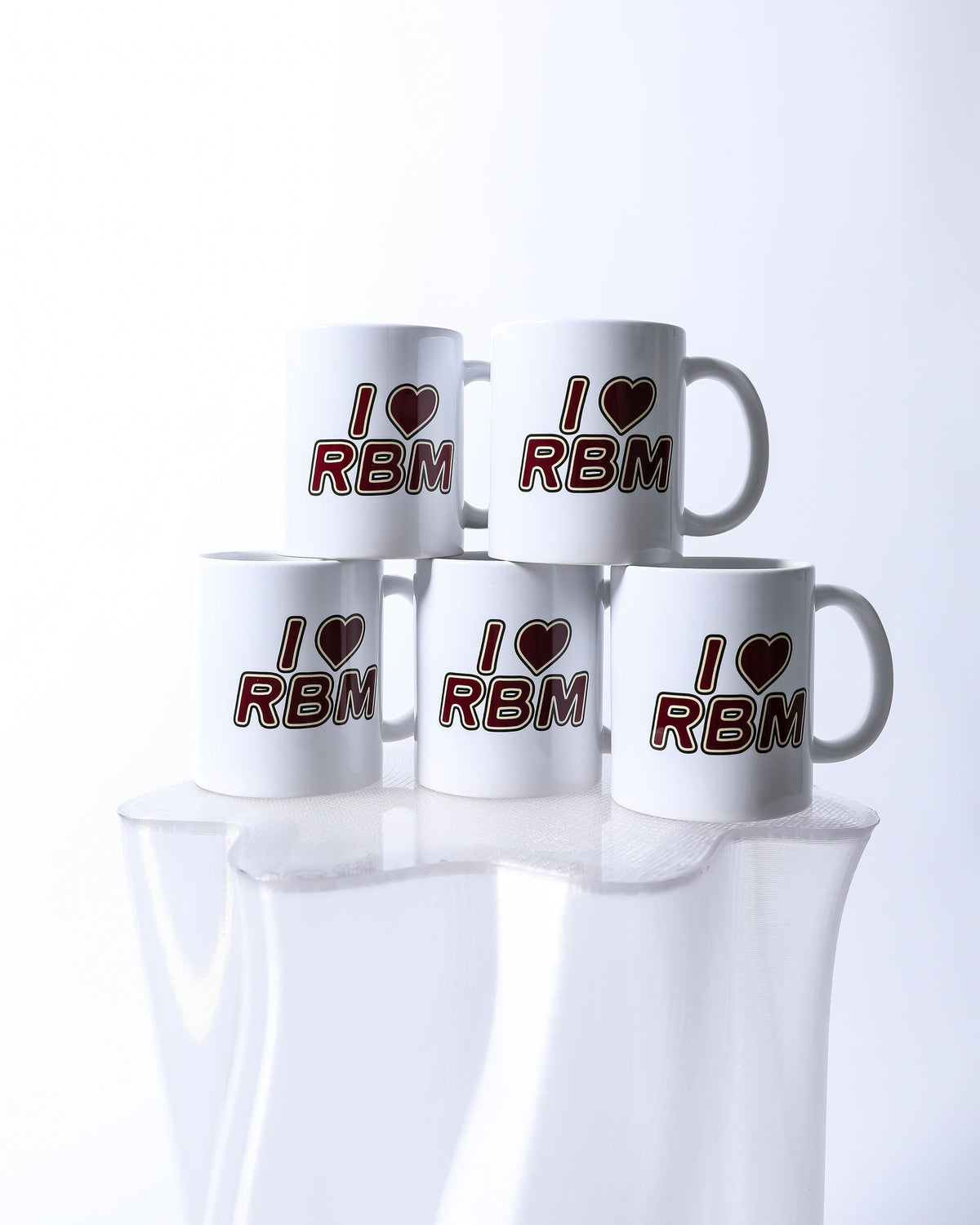 Five white mugs with 'I ❤️ RBM' text on a reflective surface.